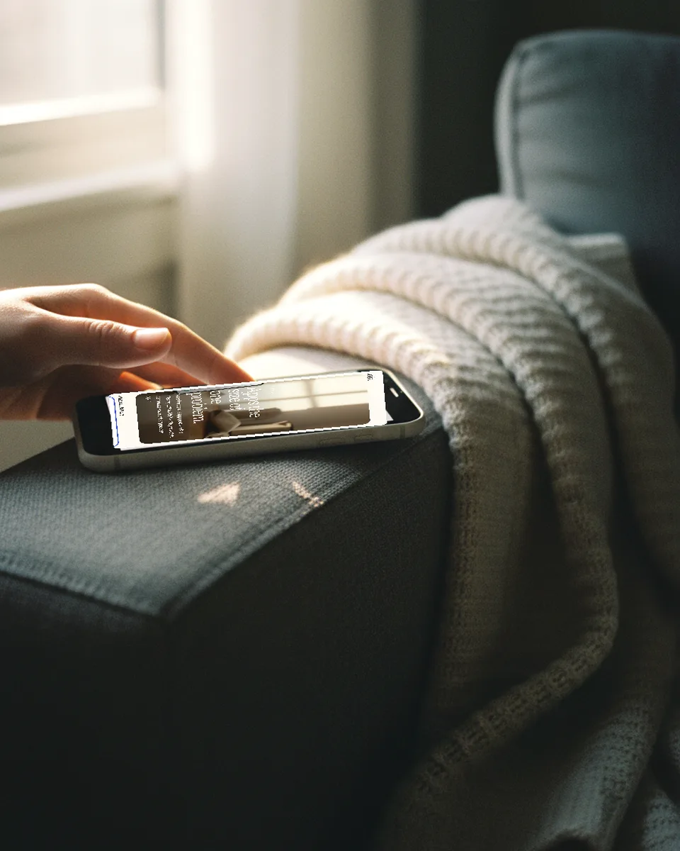 A thumb typing a sentence into the Loft prompt field on a phone, resting on a couch arm under a knit blanket in morning light.